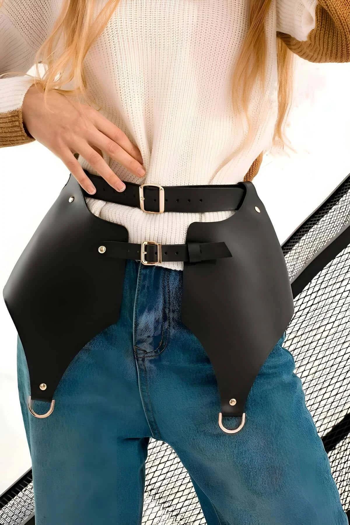 Black faux leather skirt harness belt with two gold buckle waist straps and D-ring panels worn over blue jeans