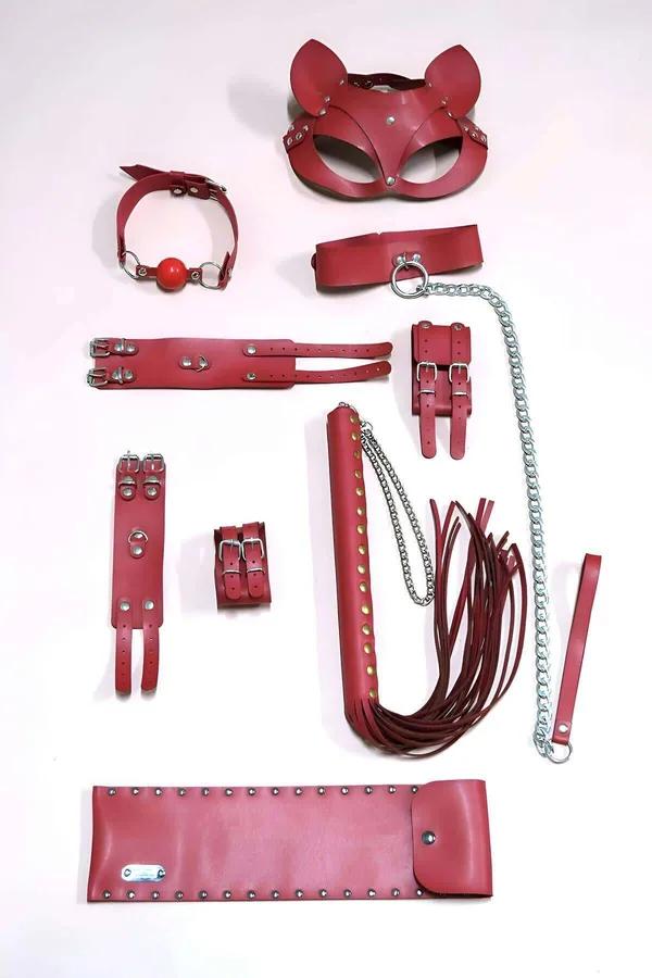 Crimson vegan leather 8-piece accessory collection flatlay with cat mask, collar, cuffs, flogger, and studded belt, silver ha