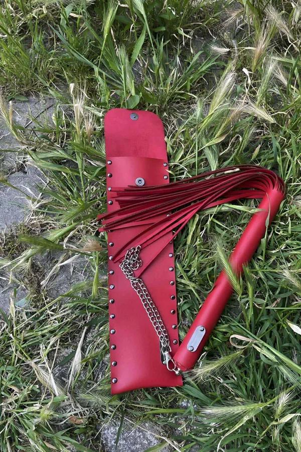 Red faux leather fashion flail with silver chain handle and fringe tails laid on grass next to studded carry pouch