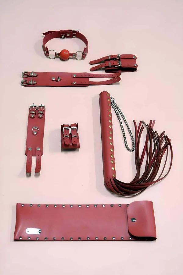 Crimson faux leather 6-piece restraint kit with wrist cuffs, ankle cuffs, ball gag, flogger, studded arm cuff, and pouch on p