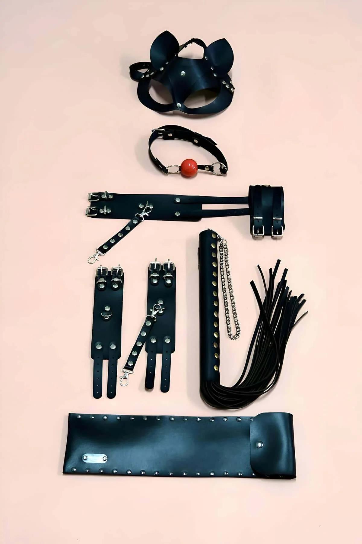 Black faux leather costume accessory set with cat mask, ball gag, spreader bar cuffs, flogger, and wide studded cuff on pink