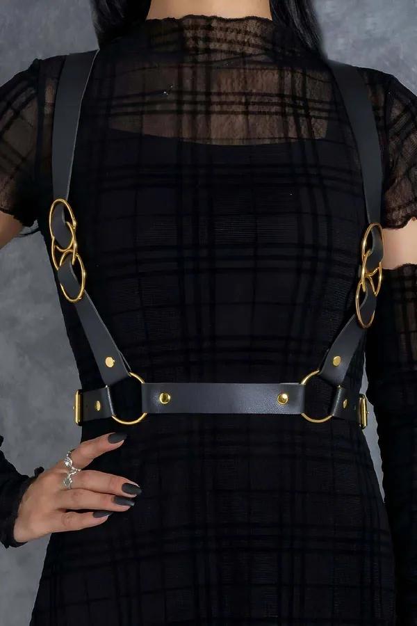 Black faux leather body harness with paired interlocking gold O-rings and adjustable waist belt worn over sheer black top