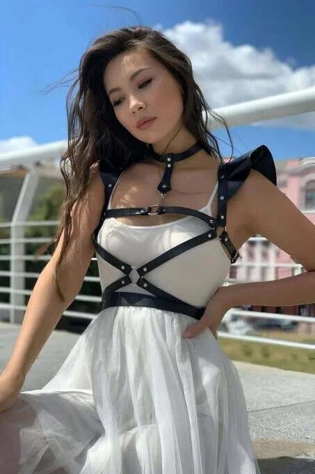 black faux leather strappy body harness with central O-ring and structured shoulder caps worn over white dress