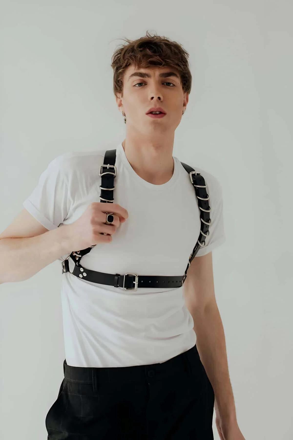 Black faux leather chest harness with silver O-rings on shoulder straps and rectangular buckle waist band worn over white t-s